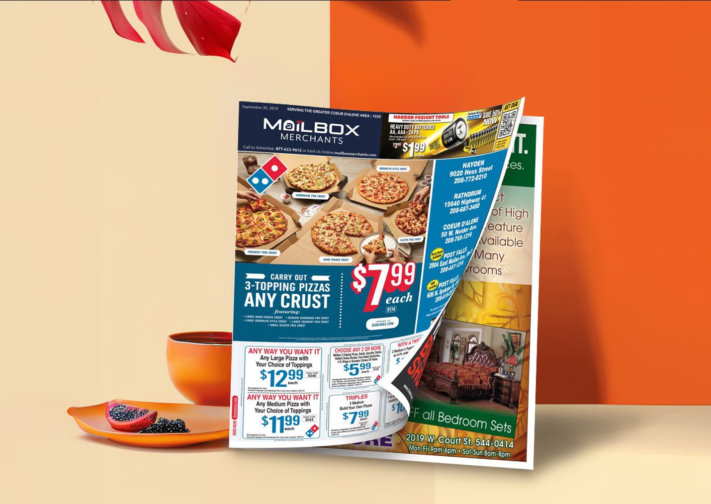 Folded direct mail advertisement from Mailbox Merchants featuring local deals, including pizza promotions and retail ads, displayed on a tabletop with a cup and plate.